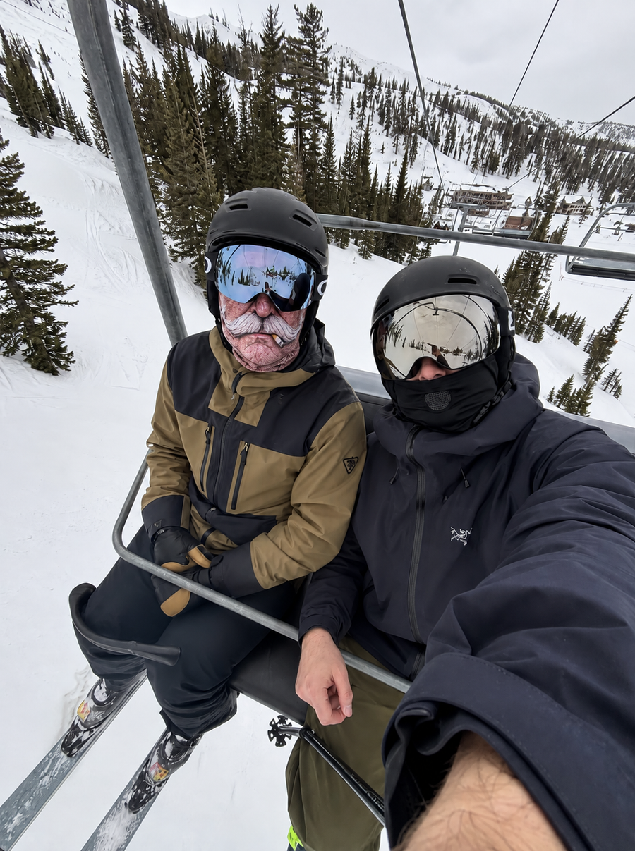 Two riders on a chairlift, one wearing a white-moustache ski mask